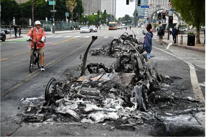 La gente observa los restos carbonizados de un vehículo Waymo incendiado en la calle tras una noche de protestas en respuesta a las operaciones federales de inmigración en Los Ángeles, el 9 de junio de 2025. (Ronaldo Schemidt/AFP vía Getty Images)