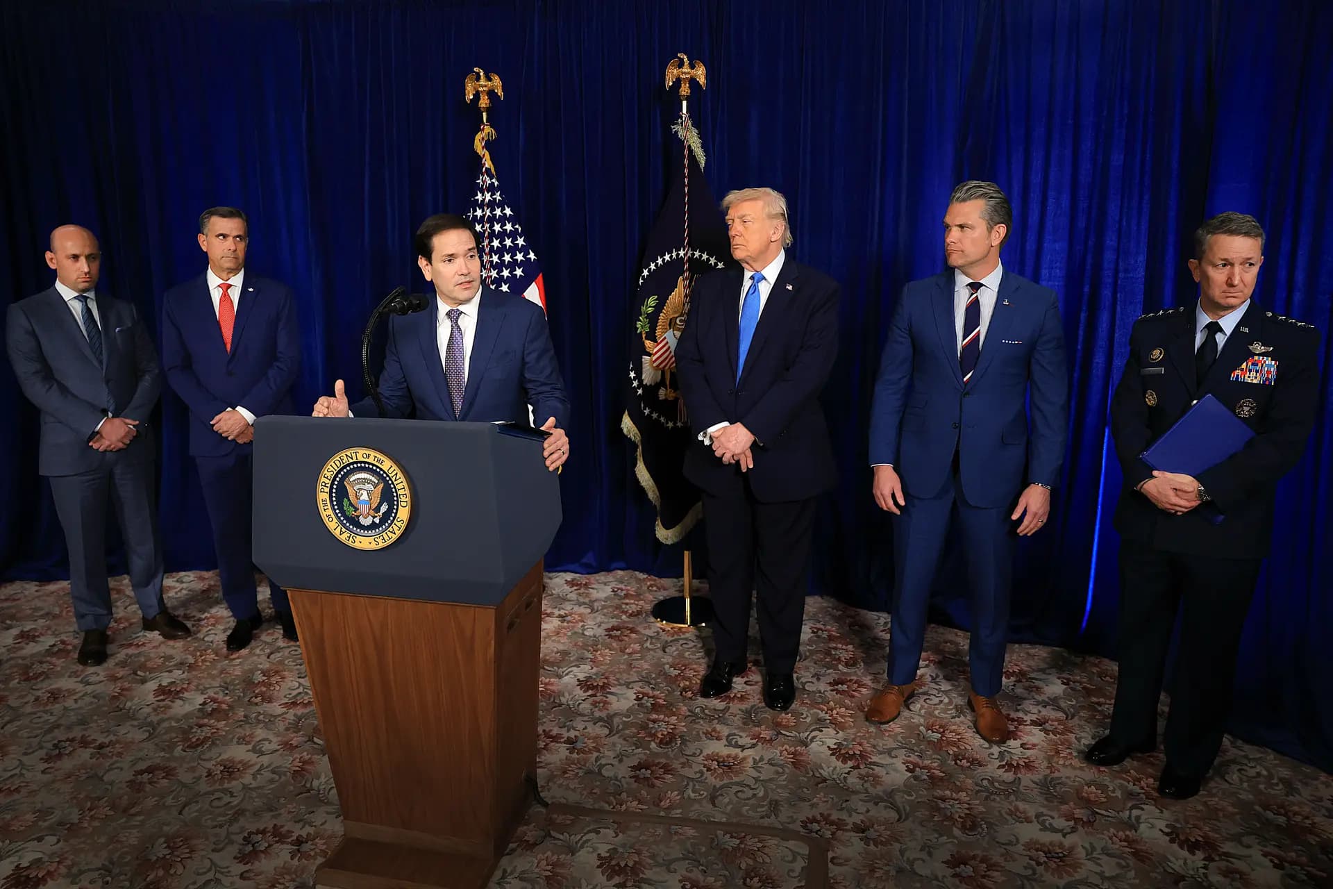 <em>El secretario de Estado, Marco Rubio (al frente), habla durante una conferencia de prensa mientras (de izq. a der.) el asesor de Seguridad Nacional Stephen Miller; el director de la CIA John Ratcliffe, el presidente Donald Trump, el secretario de Guerra Pete Hegseth y el jefe del Estado Mayor Conjunto, general Dan Caine, escuchan en el club Mar-a-Lago en Palm Beach, Florida, el 3 de enero de 2026. (Joe Raedle/Getty Images)</em>
