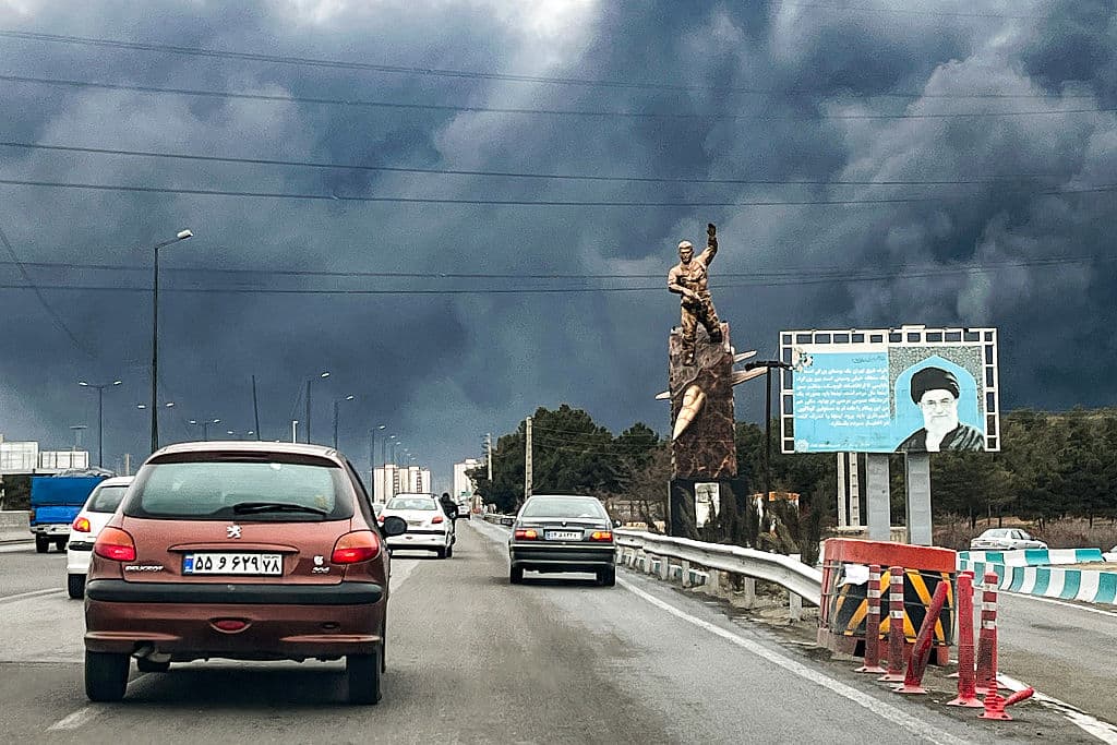 Vehículos circulan por una autopista pasando junto a una estatua conmemorativa de guerra y una valla publicitaria que muestra al difunto líder supremo de Irán, el ayatolá Alí Jamenei, que murió en un ataque aéreo el 28 de febrero, con columnas de humo negro elevándose, en Teherán, el 8 de marzo de 2026.  (Foto de AFP vía Getty Images) 