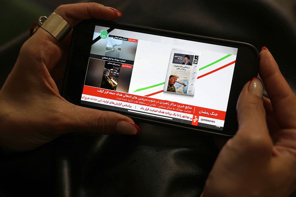 <em>Una mujer iraní ve un noticiero en un canal de televisión estatal iraní en su teléfono móvil en Teherán el 25 de marzo de 2026. Los periódicos iraníes ridiculizaron el 25 de marzo lo que llamaron las "mentiras" de Donald Trump sobre las conversaciones diplomáticas en curso para poner fin a la guerra, publicando caricaturas del presidente estadounidense al estilo de Pinocho. (Foto de AFP vía Getty Images)</em>