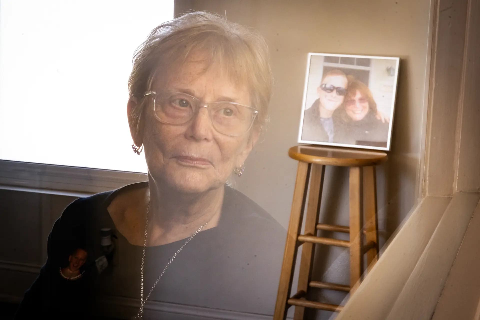Judy Rogg se refleja en una ventana con una foto de ella y su difunto hijo Erik al fondo en Los Ángeles el 15 de abril de 2026. Rogg espera que las demandas y la legislación eventualmente conduzcan a cambios, pero cree que la educación es el eslabón perdido. ( John Fredricks/The Epoch Times)