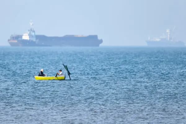 Dos hombres sentados en una pequeña embarcación en el agua, con buques de carga anclados al fondo, en el estrecho de Ormuz, frente a Bandar Abbas (Irán), el 4 de mayo de 2026. (Amirhosein Khorgooi/ISNA vía AP)