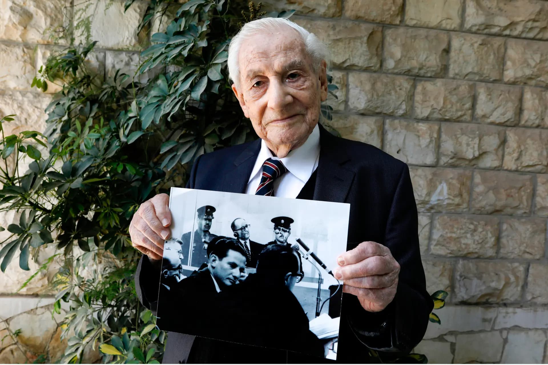 Gabriel Bach, de 93 años, exjuez del Tribunal Supremo de Israel y exfiscal adjunto durante el juicio del alto funcionario nazi alemán Adolf Eichmann, posa para una fotografía sosteniendo una imagen que lo muestra (abajo) durante el juicio de Eichmann (arriba), en el patio de su casa en Jerusalén el 3 de mayo de 2020. (Menahem Kahana/AFP vía Getty Images)