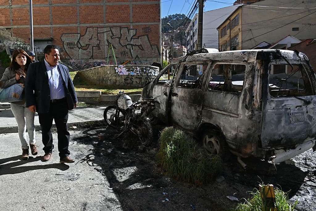<em>Varias personas observan un coche patrulla y una motocicleta calcinados en La Paz, el 19 de mayo de 2026, un día después de una protesta que exigía la renuncia del presidente boliviano Rodrigo Paz. Las protestas en Bolivia, que pedían la dimisión del presidente de centroderecha Rodrigo Paz, se intensificaron el 18 de mayo de 2026, cuando los manifestantes irrumpieron en edificios gubernamentales y un líder de las protestas enfrentó cargos de terrorismo por los disturbios. La policía antidisturbios se enfrentó a los manifestantes durante horas, el gas lacrimógeno cubrió las calles de La Paz, los comercios cerraron sus puertas y los suministros escasearon debido a los bloqueos de las protestas que obstruían las rutas de acceso a la capital. (Foto de AIZAR RALDES / AFP vía Getty Images)</em>