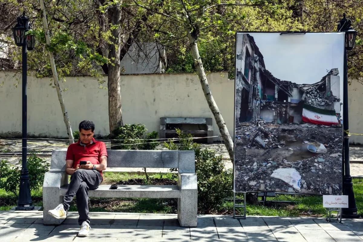 Un hombre iraní mira su teléfono móvil mientras está sentado cerca de una gran fotografía que representa la actual “guerra de Medio Oriente” en un parque de Teherán el 16 de abril de 2026. (AFP vía Getty Images)