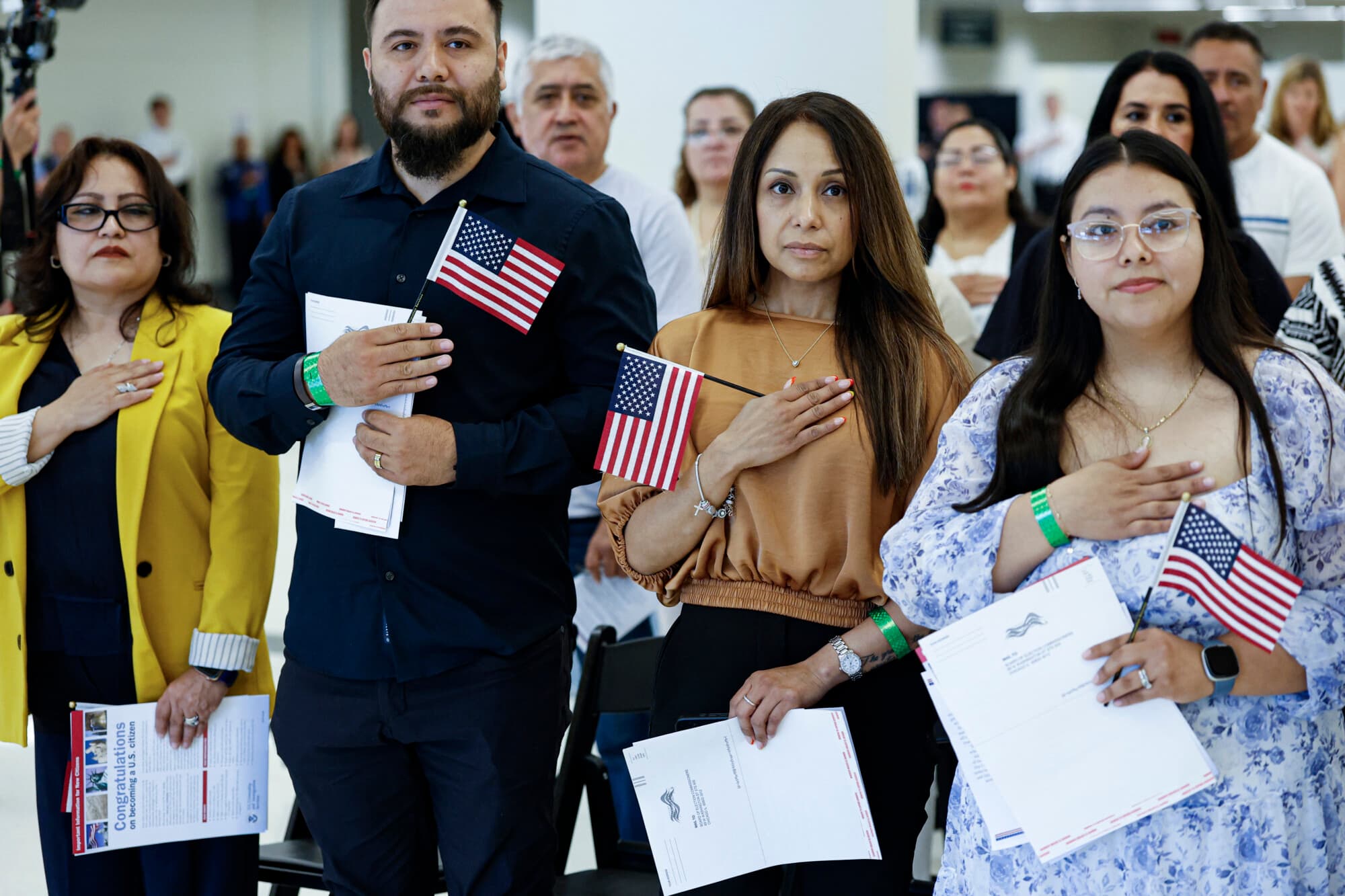 Nuevos ciudadanos estadounidenses se ponen en pie para escuchar el himno nacional antes de prestar el juramento de lealtad y recibir sus certificados de naturalización durante una ceremonia formal en el Aeropuerto Internacional Midway de Chicago, Illinois, el 25 de junio de 2025. (KAMIL KRZACZYNSKI/AFP vía Getty Images)