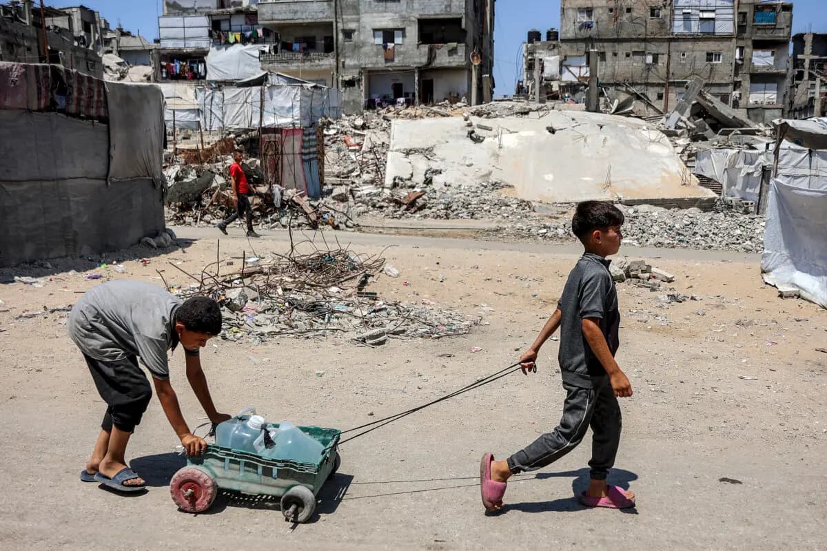 <em>Unos niños transportan un carro cargado de bidones de agua pasando junto a tiendas de campaña y escombros en un campamento para personas desplazadas por la guerra en el barrio de Sheikh Radwan, en la ciudad de Gaza, el 6 de julio de 2026. (Omar Al-qattaa /AFP vía Getty Images).</em>