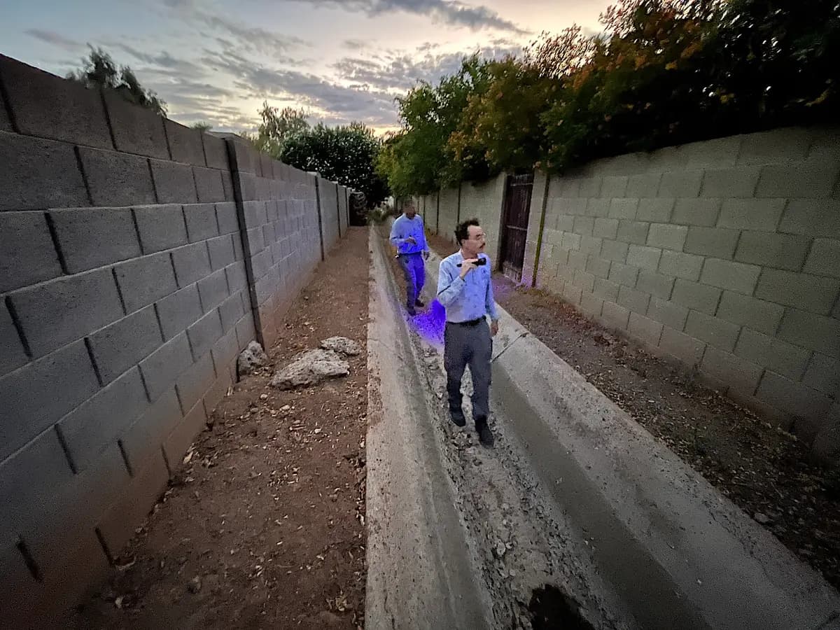 Jeremiah Sourp (derecha) y Mike Golleher caminan en busca de escorpiones a lo largo del canal de drenaje de concreto detrás de un barrio residencial en Tempe, Arizona, el 24 de junio de 2026. (Allan Stein/The Epoch Times)