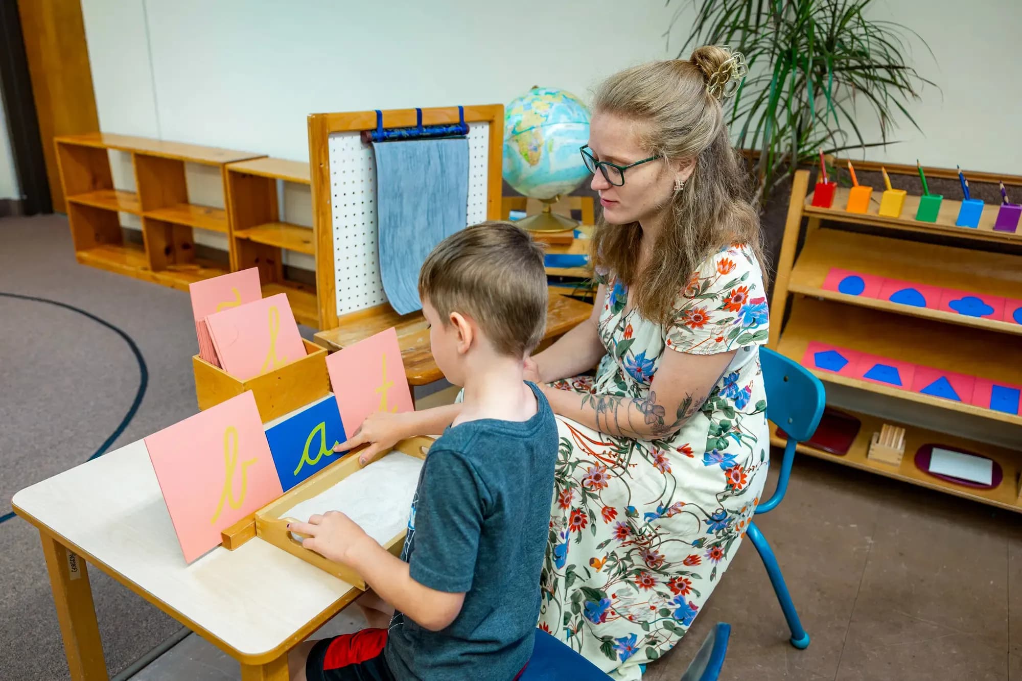 La maestra Colleen Endrizzi observa a un alumno de primer grado trazar letras cursivas en una bandeja de arena, en la escuela Willoughby Montessori en Willoughby, Ohio, el 10 de julio de 2026. (Jonathan D. Williams para The Epoch Times).