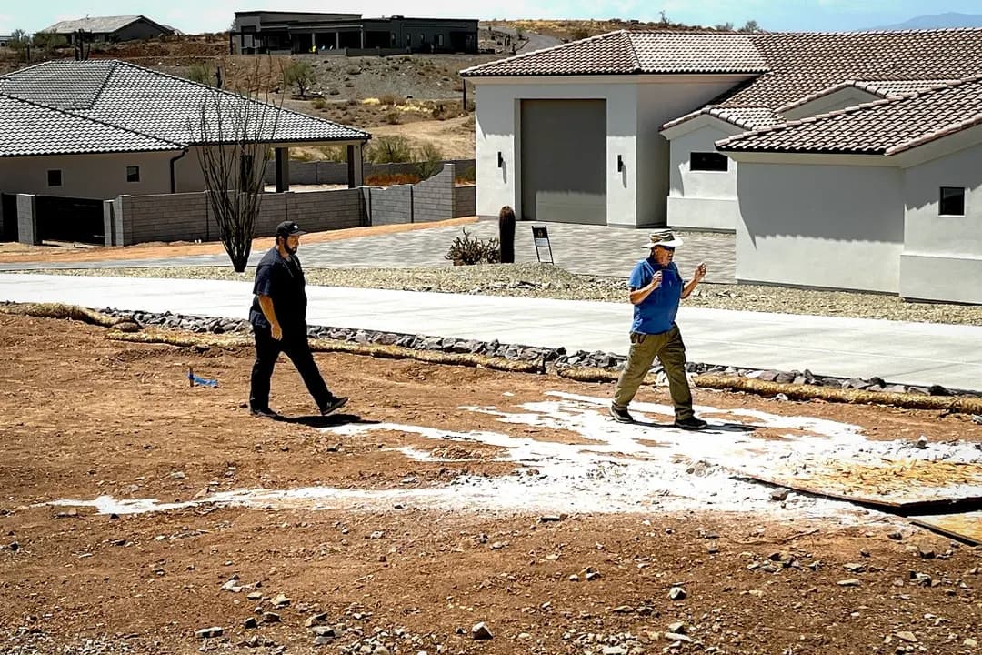 El gerente de construcción de Diamante Homes, Tanner Cook (izquierda), sigue a Jack Coel mientras este busca agua subterránea utilizando la antigua práctica de la radiestesia en Phoenix, el 13 de julio de 2026. Cook recurrió a este método ancestral para encontrar agua cuando los pozos que abastecían a un desarrollo en construcción comenzaron a secarse. (Allan Stein/The Epoch Times)