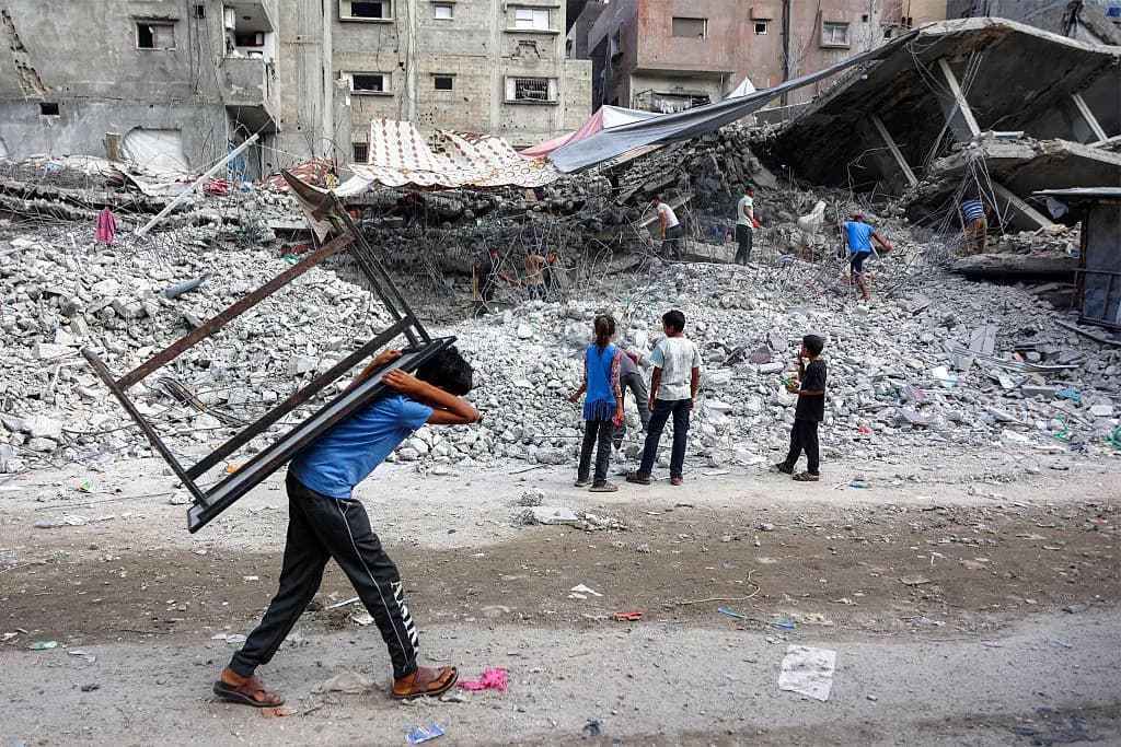 Un niño que lleva una mesa a la espalda pasa junto a trabajadores de rescate que retiran los escombros de un edificio destruido en la ciudad de Gaza el 16 de agosto de 2026. (Omar Al-Qattaa/AFP vía Getty Images)