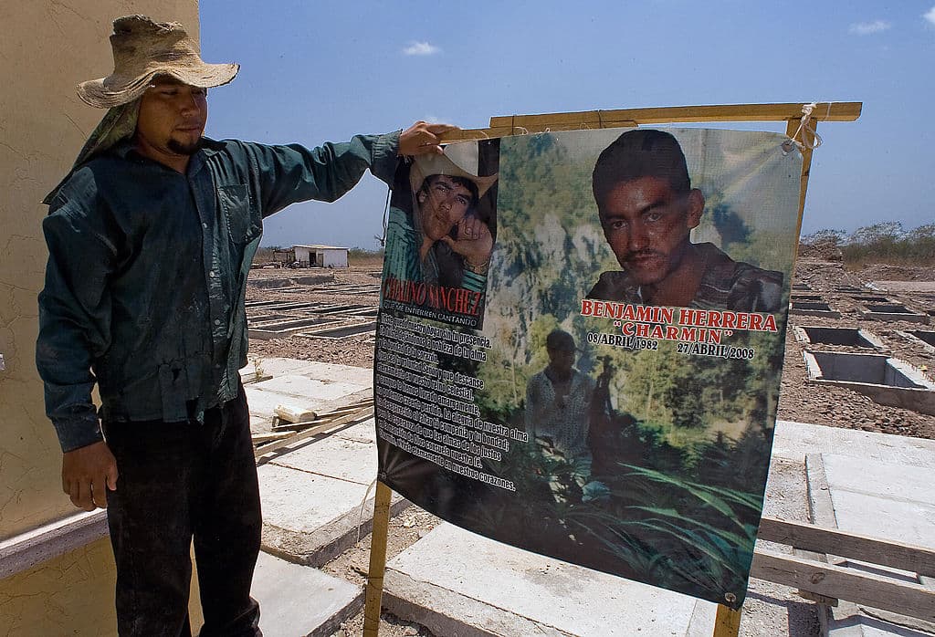 <em>Trabajador del cementerio de Culiacán, Sinaloa, sostiene un cartel mientras relata la historia de un presunto sicario, el 3 de junio de 2008. México atravesaba una guerra entre cárteles por el control territorial y las rutas de tráfico hacia Estados Unidos, marcada por la violencia extrema, ejecuciones, decapitaciones, torturas y amenazas contra grupos rivales y las fuerzas de seguridad. En ese año se habían registrado al menos 1378 muertes, un 47 % más que en el mismo periodo de 2007. (Luis Acosta / AFP vía Getty Images)</em>