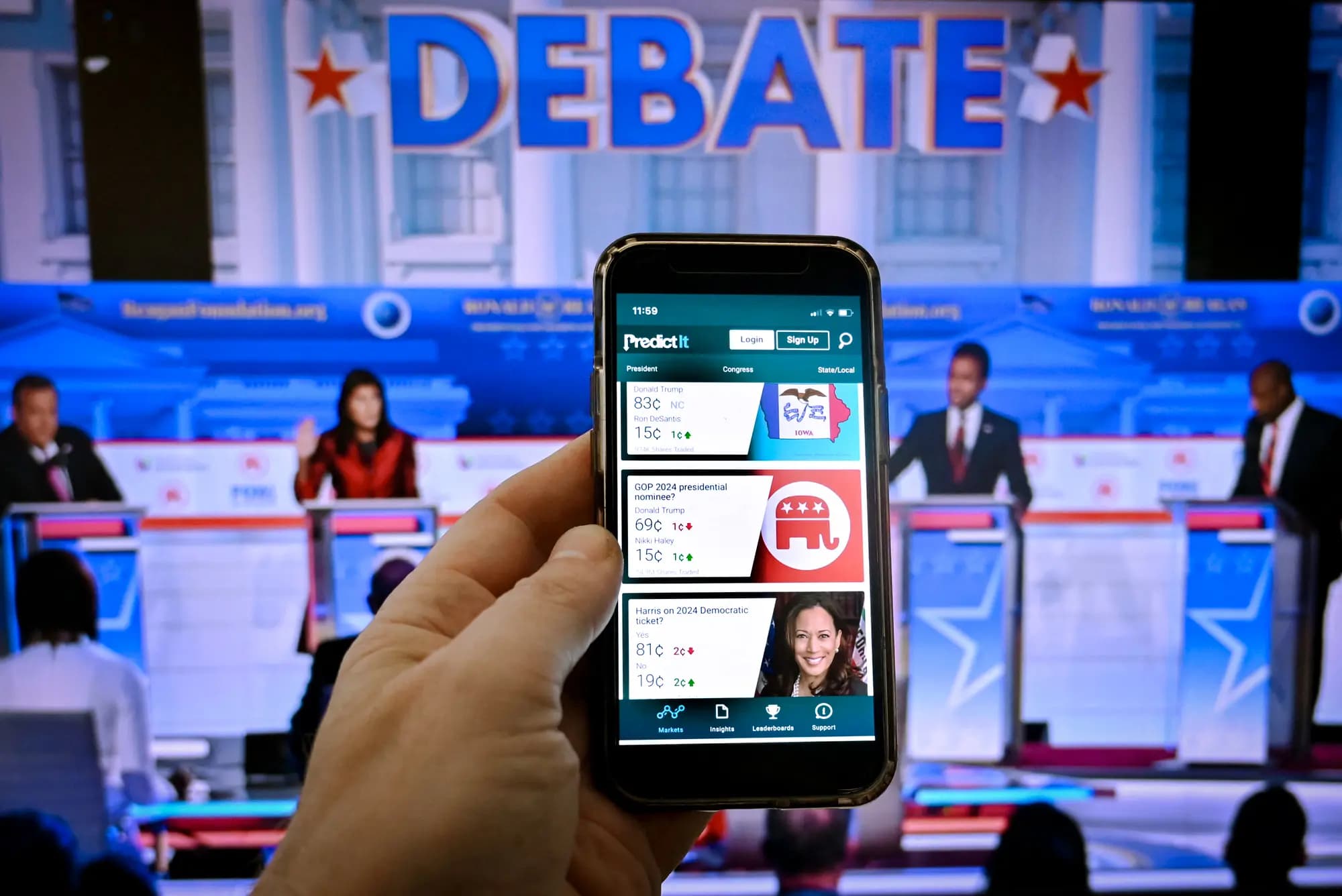 <em>Esta ilustración fotográfica, tomada en Washington el 1 de noviembre de 2023, muestra la pantalla de un teléfono con la plataforma de predicción en línea PredictIt y una imagen del segundo debate de las primarias presidenciales republicanas en Simi Valley, California, el 27 de septiembre de 2023. Los votantes, en busca de información y contexto sobre las contiendas electorales clave, consultan cada vez más los informes de los mercados de predicción antes de emitir su voto. (Olivier Douliery/AFP vía Getty Images).</em>