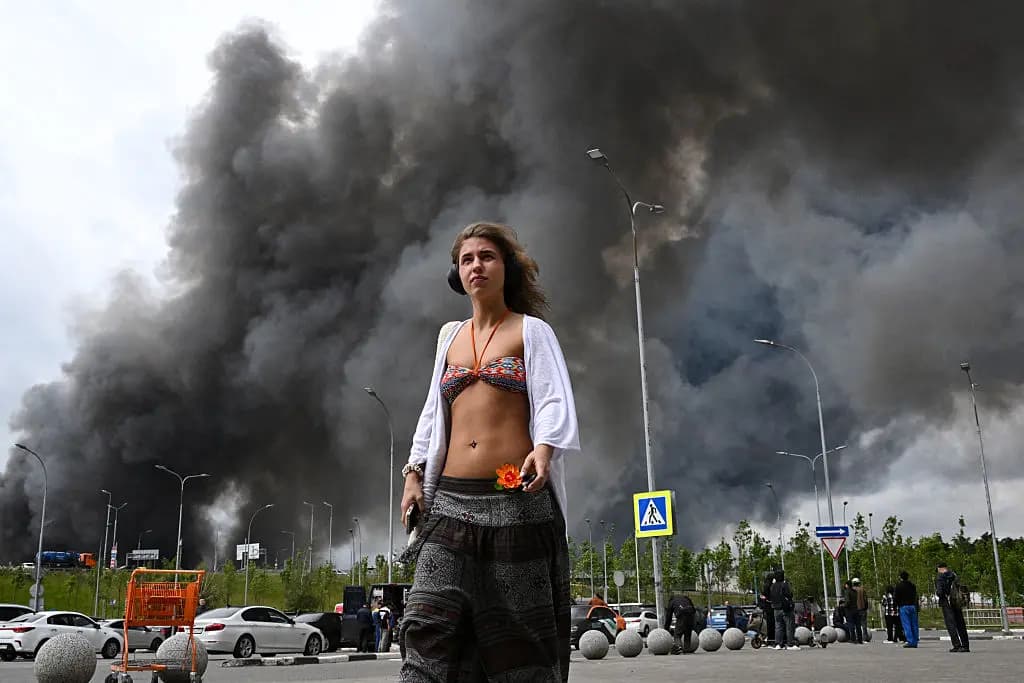Una mujer camina frente a un centro comercial mientras se eleva humo negro desde la zona de la refinería de la empresa petrolera rusa Gazprom Neft, ubicada en las afueras del sureste de Moscú, el 18 de junio de 2026. (AFP vía Getty Images).