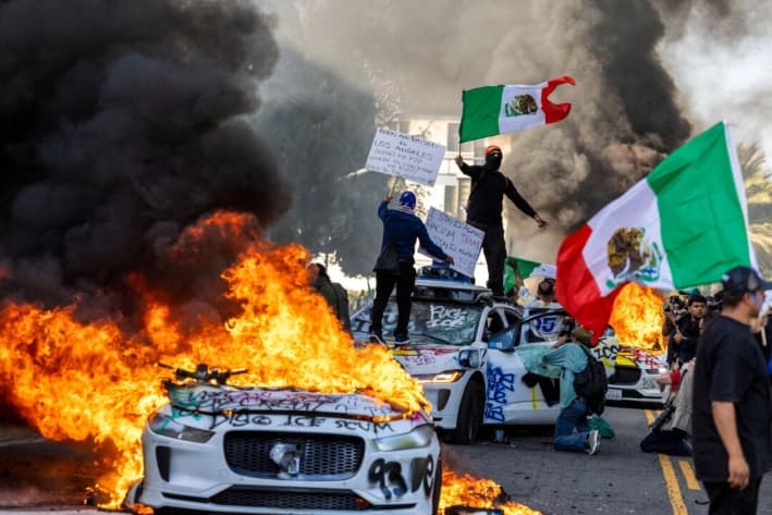 Manifestantes ondean banderas desde lo alto de un coche destrozado, junto a vehículos de Waymo en llamas, mientras se enfrentan con las fuerzas del orden en las calles cercanas al edificio federal durante una protesta contra las operaciones federales de inmigración, en Los Ángeles, el 8 de junio de 2025. (Ringo Chiu/AFP vía Getty Images)